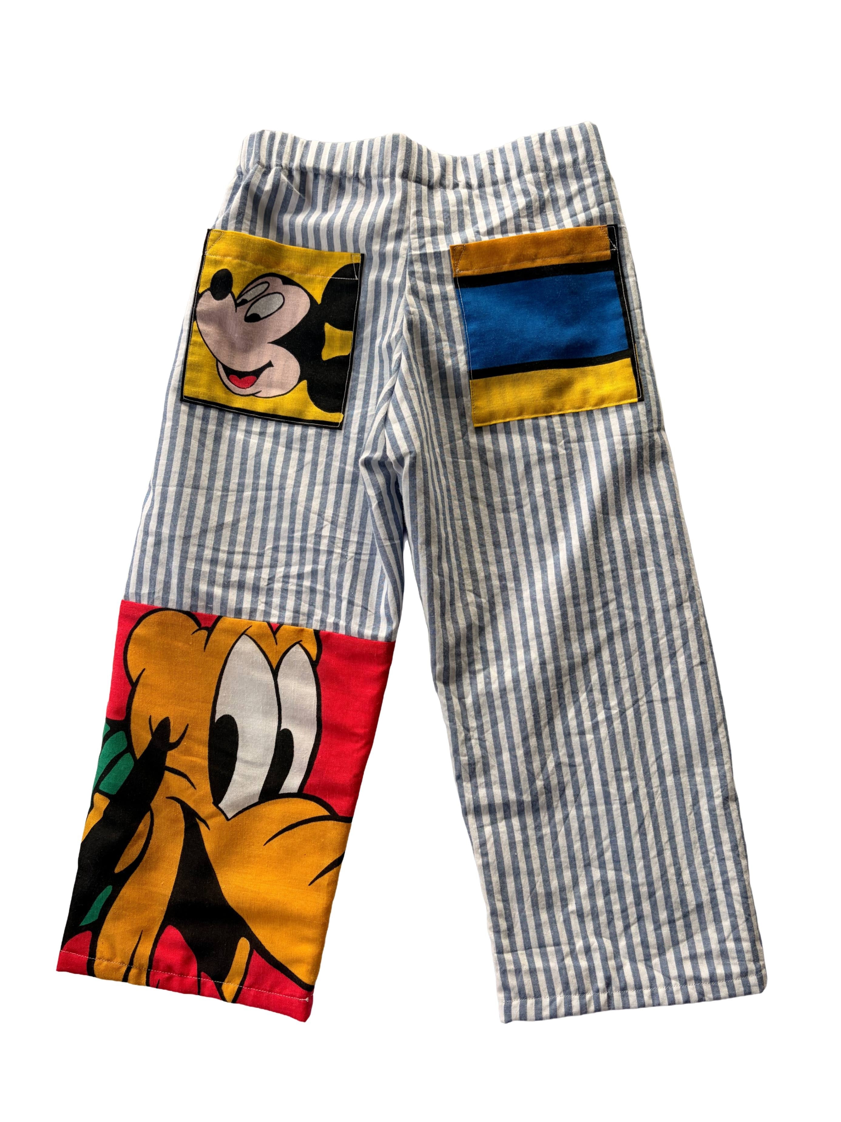 A pair of children's pants featuring a blue and white vertical stripe pattern, with colourful cartoon character patches on the back pockets. mickey mouse pocket and pluto at the bottom of the pant leg.